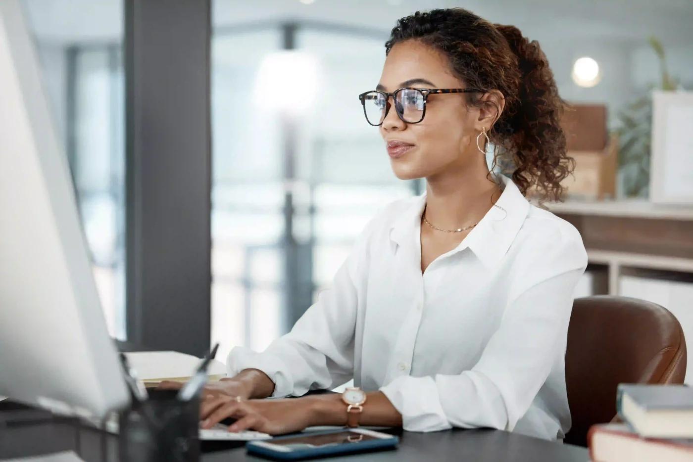 Woman wearing glasses and white shirt working on a computer in the Tasklaw system in a modern and bright office.